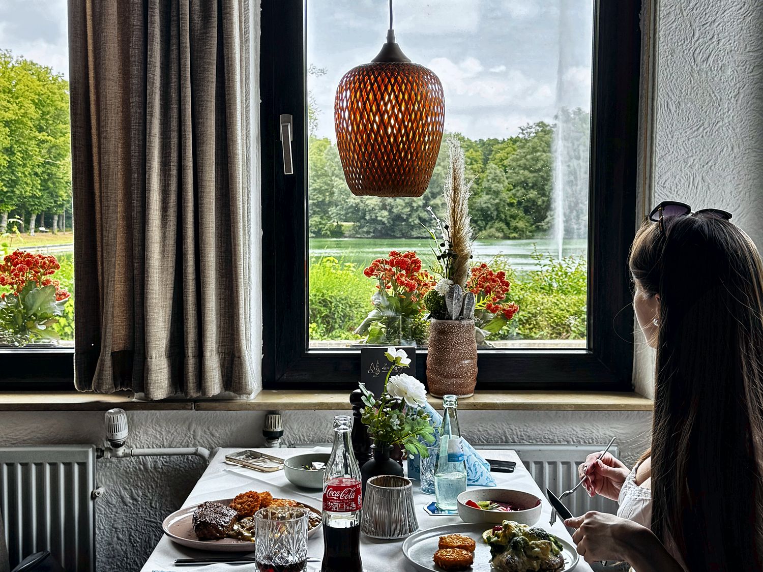 Ein gedeckter Tisch am Fenster mit zwei Tellern vom Grill, dahinter der Blick über den Teich zur Fontäne